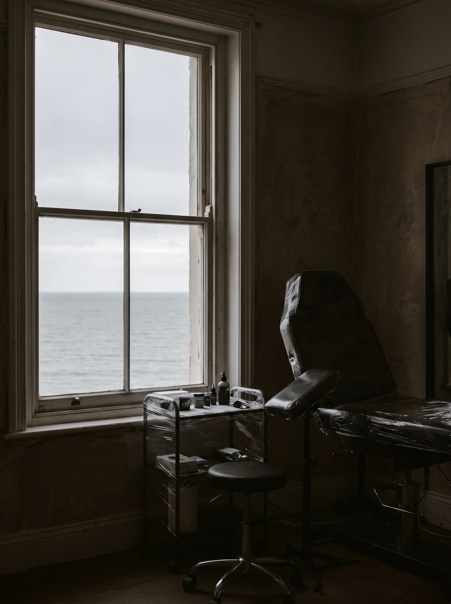Tall studio window with the flat grey Margate horizon beyond, a corner of a wrapped tattoo work station visible in shadow
